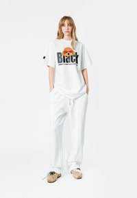 Camisa blanca de gran tamaño con un diseño gráfico que presenta una galleta y texto. Combinada con pantalones blancos de corte relajado y zapatillas beige.