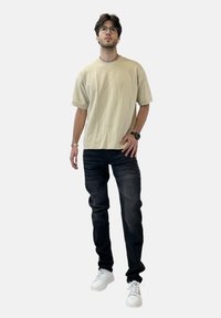 Camiseta de algodón color beige claro con mangas cortas, combinada con jeans oscuros y zapatillas blancas, de pie contra un fondo neutro.