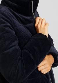 Person som drar upp dragkedjan på en mörkblå kordflorsjacka med hög krage, visar händer och detaljer av den texturerade tyget.