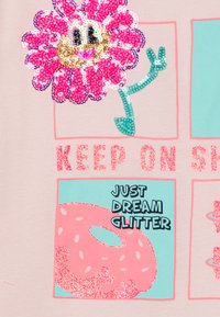 Ljust rosa skjorta med en paljetterad blomma, texten "KEEP ON SH" i glitter och en donutgrafik med glittriga detaljer och texten "JUST DREAM GLITTER."