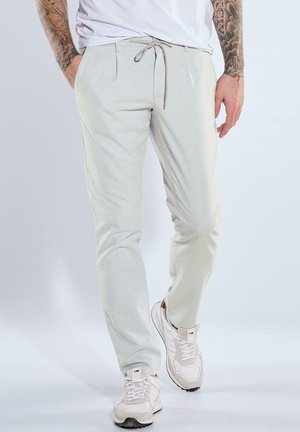 Mann trägt hellbeige Kordelzughose und weiße Turnschuhe, steht mit den Händen in den Taschen vor einem schlichten Hintergrund.