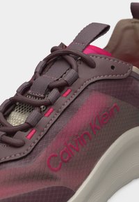 Κοντινό πλάνο μοβ και ροζ αθλητικού παπουτσιού Calvin Klein με υφασμένα κορδόνια και λευκή σόλα, που αναδεικνύει το λογότυπο στην πλαϊνή πλευρά.