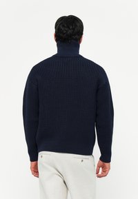 Marineblauwe gebreide trui met een hoge kraag, met een gestructureerd patroon en geribbelde manchetten, gezien vanaf de achterkant tegen een witte achtergrond.