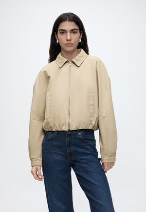 Mujer con el pelo largo y oscuro, vestida con una chaqueta beige con cremallera y vaqueros azul oscuro, de pie frente a un fondo claro liso.