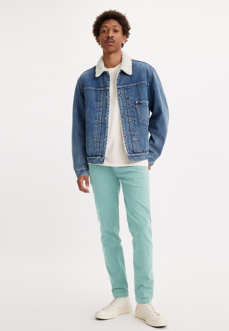 Chaqueta de denim con un cuello de borreguito blanco, de color azul desgastado, con botones y un bolsillo. Llevada sobre una camisa blanca y pantalones verde menta.