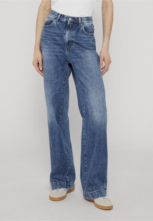 Personne portant un jean bleu taille haute à jambes larges et des baskets blanches, debout les mains détendues le long du corps.