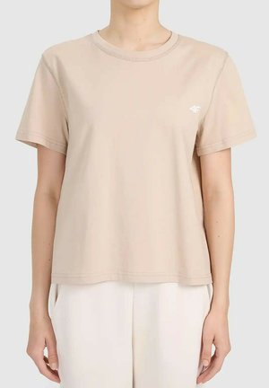 Beżowy t-shirt z krótkim rękawem i okrągłym dekoltem na osobie w białych spodniach, małe białe logo na lewej piersi, neutralne tło.