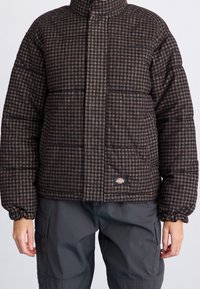 Veste rembourrée à carreaux marron avec texture matelassée, col montant et poignets élastiques. Comprend un patch logo près de l'ourlet.