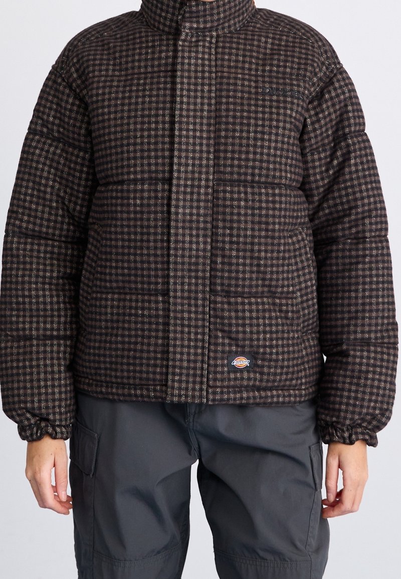 Veste rembourrée à carreaux marron avec texture matelassée, col montant et poignets élastiques. Comprend un patch logo près de l'ourlet.