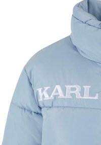 Lys blå pufferjakke med høj krave, der har hvid broderet tekst med "KARL" på forsiden. Glat, blank overflade.