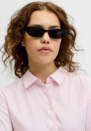 Młoda kobieta o kręconych brązowych włosach, nosząca czarne okulary przeciwsłoneczne typu cat-eye oraz białą koszulę z cienkimi czerwonymi pionowymi paskami, z neutralnym wyrazem twarzy.