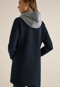 Femme portant un sweat à capuche noir à manches longues avec une capuche grise, vue de dos, mains dans les poches, fond neutre.