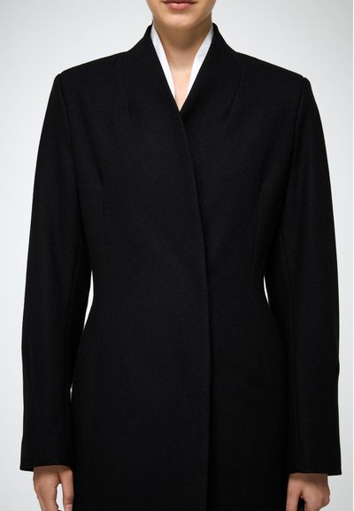 Cappotto di lana nera con colletto alto, maniche lunghe e silhouette aderente. Tessuto liscio con linee pulite e senza chiusure visibili.
