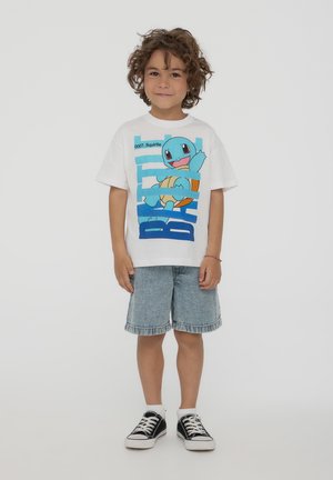 Maglietta bianca in cotone con grafica di Squirtle blu, colletto rotondo, maniche corte, abbinata a pantaloni corti in denim chiaro e sneaker nere.