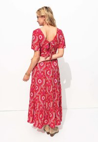 Robe maxi à motif floral rouge avec des manches courtes, un design à nouer dans le dos et un ourlet fluide. Le motif présente des éléments paisley et des motifs de soleil.