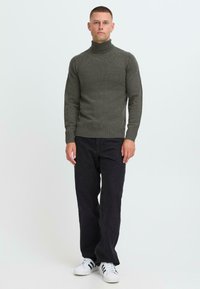 Dunkelgrüner Rollkragenpullover mit gerippter Textur, kombiniert mit lockeren schwarzen Hosen. Das Model trägt weiße Sneaker mit schwarzen Streifen.