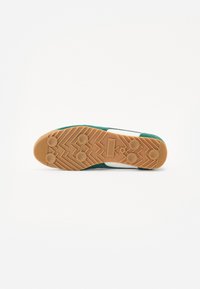 Grüne Sneaker mit einer Gummisohle, die ein strukturiertes Grip-Muster und weiße Akzente aufweist. Das Obermaterial wirkt weich und suedeartig.