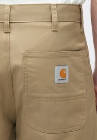 Παντελόνι εργασίας σε καμηλό χρώμα με πίσω τσέπη που φέρει λευκή ετικέτα με πορτοκαλί λογότυπο. Το ύφασμα έχει μια διακριτική υφή.