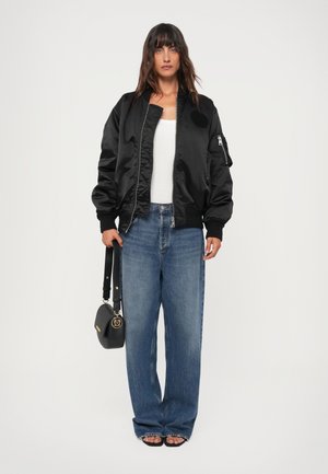 Black satin bomber jacket with ribbed cuffs, zip closure, and side pocket. Light-colored tank top, loose blue jeans, and black handbag.