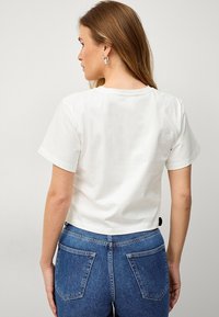 T-shirt bianco a maniche corte in cotone con scollatura a girocollo, caratterizzato da un design cropped e un'etichetta nera vicino all'orlo.