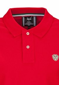 Rode polo met een kraag, twee witte knopen en een schildlogo op de linkerborst. Het materiaal lijkt textuur te hebben en de stof is glad.