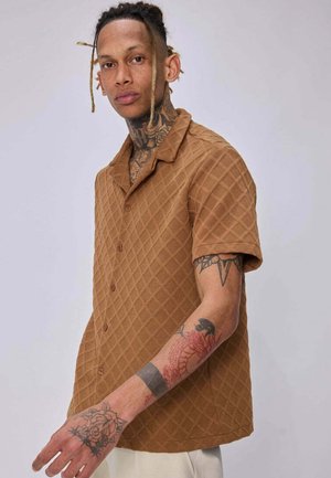 Homme avec dreadlocks et tatouages porte une chemise marron texturée à manches courtes avec boutons et un pantalon clair sur un fond uni.