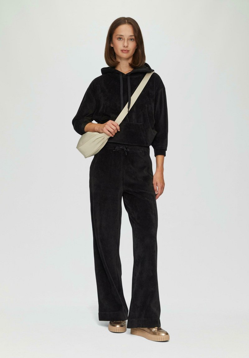 Czarny welurowy hoodie z kieszenią z przodu i kapturem, w zestawie z dopasowanymi spodniami o szerokich nogawkach. Beżowa torebka na ramię i metaliczne buty na platformie.