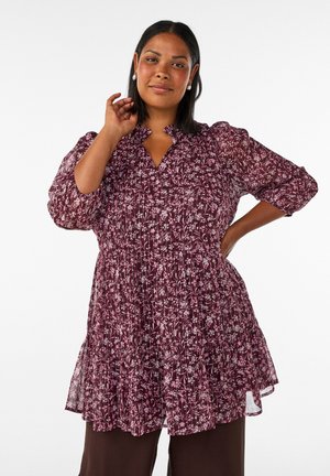 Femme debout, une main sur la hanche et l'autre touchant son visage, portant une robe fleurie violette et des boucles d'oreilles sur fond blanc.