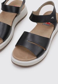 Sandalias de cuero negro con dos correas anchas, aseguradas por una correa de tobillo. Presenta una plantilla acolchada beige y una suela de goma blanca.