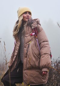 Hnedá prešívaná bunda s podšívkou s leopardím vzorom, vybavená zipsami a vreckami, nosená cez sivý sveter; doplnená žltou čiapkou a batohom.