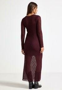 Mauve blondekjole med lange ærmer, der har et åbent vævemønster og en tætsiddende silhuet. Kjolen er semitransparent ved den nedre kant.