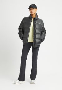 Giubbotto piumino nero con finitura lucida, maniche lunghe e colletto alto, abbinato a un top giallo chiaro e pantaloni neri. Sneakers argentate completano il look.