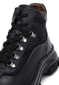 Botas negras de caña alta, hechas de cuero y fieltro. Cuentan con cordones negros, ojales metálicos y una suela texturizada para un mejor agarre.