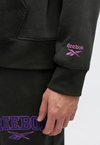 Melna svītrojuma kapuce ar ribotām aprocēm, kurai ir spilgti rozā Reebok logo uz piedurknes un violets logo uz priekšpuses. Mīksta tekstūra.