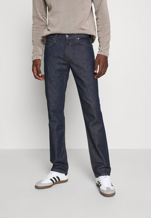 Personne portant un jean droit bleu foncé, un haut beige à manches longues et des baskets blanches avec des rayures noires, debout devant un fond uni.