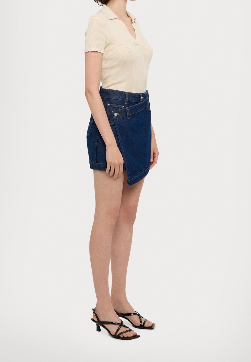 Femme portant un haut côtelé crème à manches courtes, une mini-jupe en denim sombre asymétrique et des sandales noires à lanières et talons bas, debout devant un fond blanc.