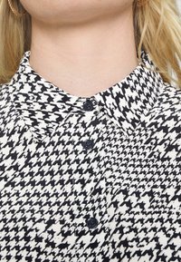 Chemise à motif pied de poule noir et blanc avec un col classique, ornée de deux boutons sombres et fabriquée dans un tissu lisse et texturé.