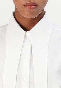 White button-up shirt with a spread collar, featuring a clean, smooth texture and a single button closure at the neck. No patterns or embellishments.