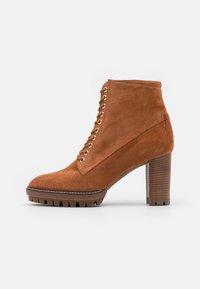 Stivaletto in suede marrone con lacci frontalmente, occhielli dorati e tacco spesso marrone. Suola in gomma testurizzata con un motivo seghettato per una migliore trazione.