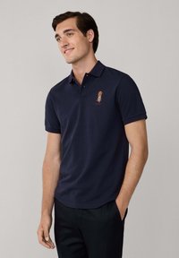 Marinblå polo med treknappsknäppning, korta ärmar och broderad logga på bröstet; tillverkad av mjukt, andningsbart material.