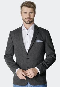 Schwarzer strukturierter Blazer mit Reverskragen, zwei Knöpfen und Fronttaschen. Tragen über einem weißen Hemd, das ein blau gemustertes Einstecktuch zeigt.