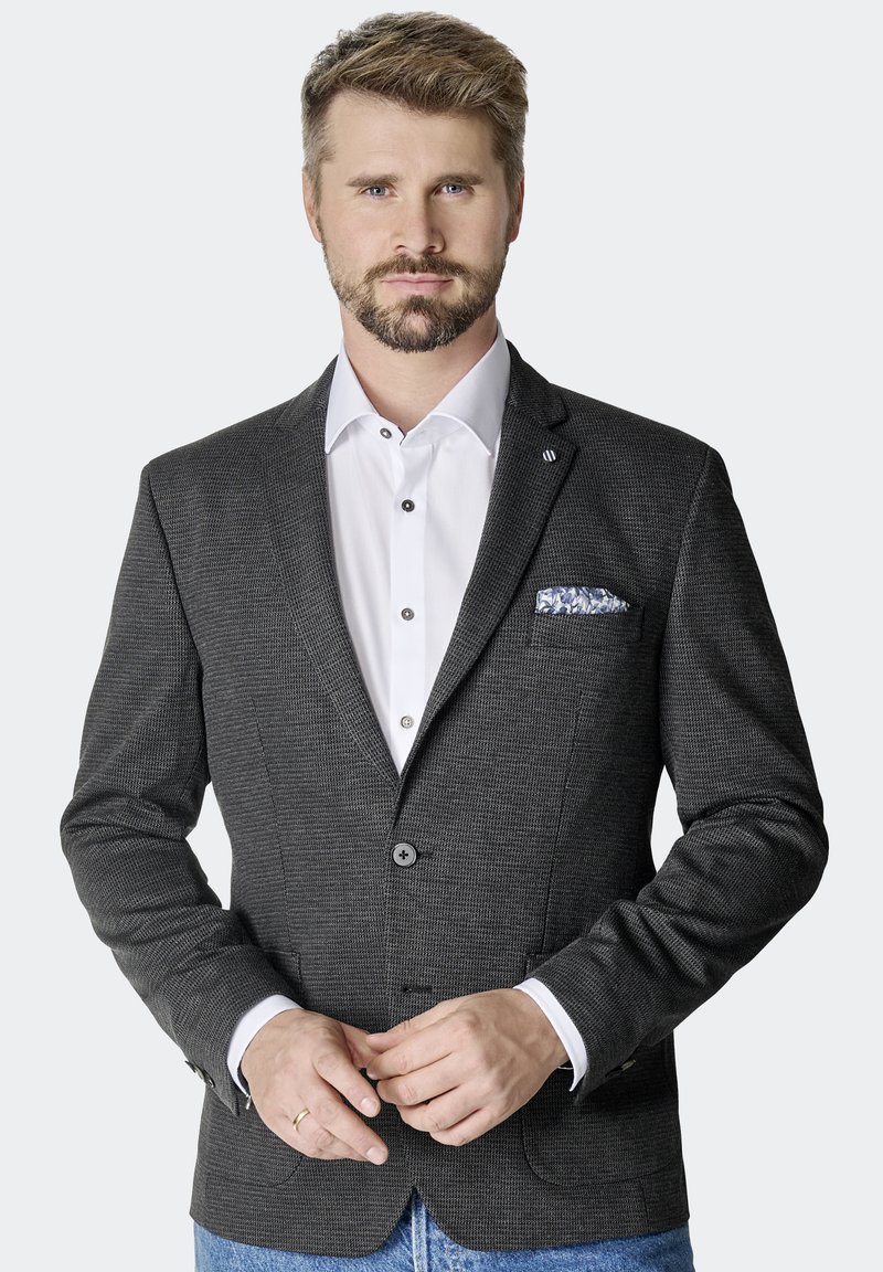 Schwarzer strukturierter Blazer mit Reverskragen, zwei Knöpfen und Fronttaschen. Tragen über einem weißen Hemd, das ein blau gemustertes Einstecktuch zeigt.