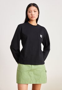 Schwarzes langärmliges T-Shirt aus Baumwolle mit einer Brusttasche, die ein Logo zeigt; kombiniert mit einem grünen Jeansrock mit Vorderentaschen.
