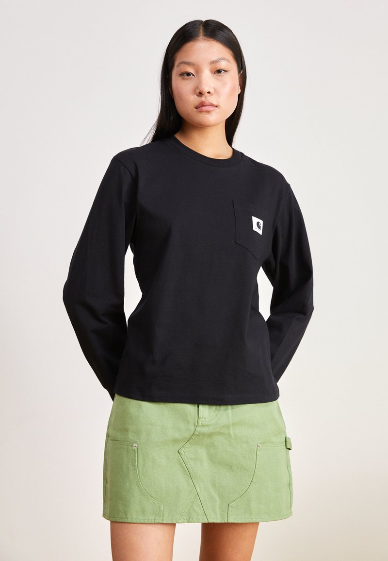 Schwarzes langärmliges T-Shirt aus Baumwolle mit einer Brusttasche, die ein Logo zeigt; kombiniert mit einem grünen Jeansrock mit Vorderentaschen.