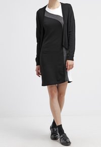 Czarna sukienka z szarymi i białymi akcentami; krótkie rękawy, dopasowana sylwetka, miękka tekstura. Połączona z krótkim czarnym kardiganem i czarnymi butami.