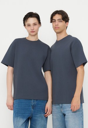 Zwei junge Erwachsene, die dunkelgraue übergroße T-Shirts und blaue Jeans tragen, stehen nebeneinander vor einem schlichten weißen Hintergrund.