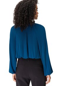 Femme aux cheveux bouclés portant un chemisier plissé à manches longues de couleur sarcelle, rentré dans un pantalon noir, de dos face à la caméra.