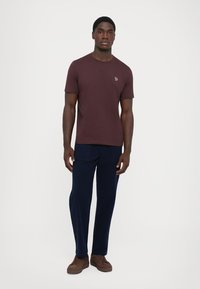 Bordeaux kortemouw T-shirt met een ronde halslijn en een klein logo op de borst, gecombineerd met marineblauwe broeken en bruine sneakers.