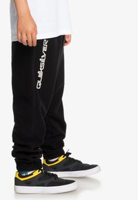 Svarta mjukisbyxor med vitt "Quiksilver"-logotyp, tillsammans med svarta skor med gula detaljer. Byxorna har en avslappnad passform och mjuk textur.