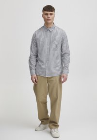 Camicia a righe a maniche lunghe bianca e blu navy, con bottone frontale, due tasche sul petto e colletto, abbinata a pantaloni beige.